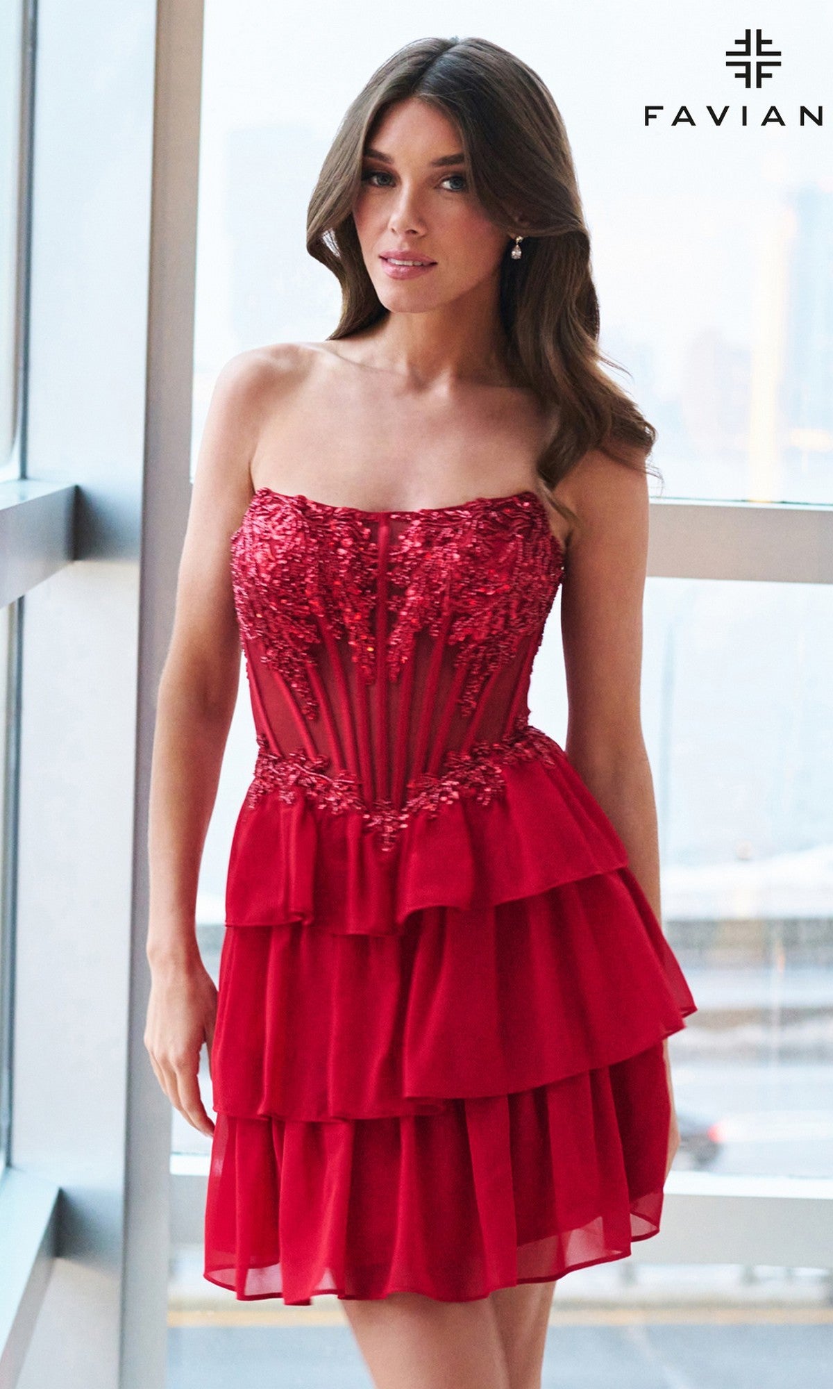 A woman in the Faviana 11500 Short Homecoming Dress—strapless, red, with lace and tiered ruffles and a corset bodice—stands indoors by large windows;